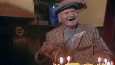 Man in a flat cap sitting in a pub with a birthday cake while holding knife.