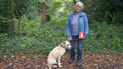 A woman with grey hair wearing a blue jacket and blue jeans stands with a light colured dog in an overgrown graveyard