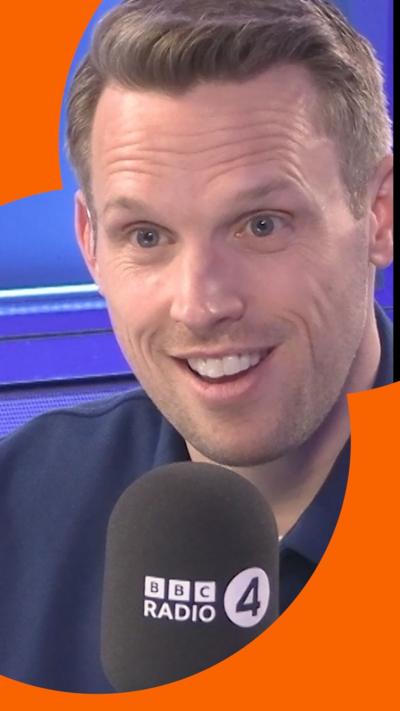 A man with short, light brown hair smiles as he talks into a microphone