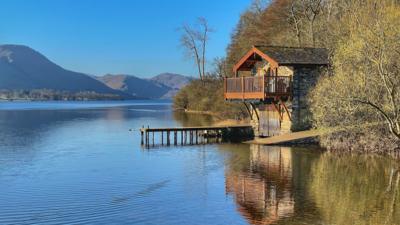 A lake with a wooden hut, two platforms into water, trees to the right, mountains in the background, under a bright blue sky