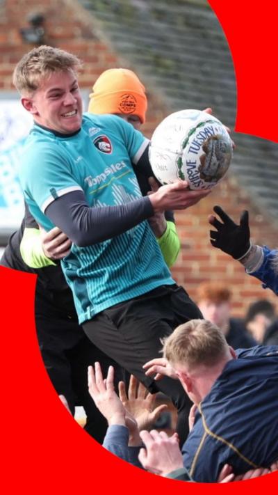 Man wearing a blue jersey and holding a white ball is standing on top of a crowd of people.