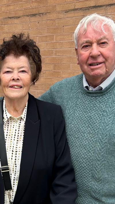 Woman with dark hair and black jacket stood next to man with grey hair and green jumper. 