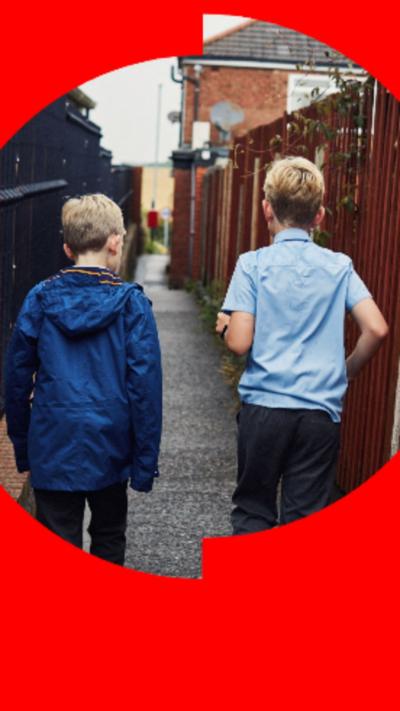 Two children playing outside seen from behind - one is wearing a blue school uniform style shirt with short sleeves, the other has a blue rain jacket on - both look to be under 10
