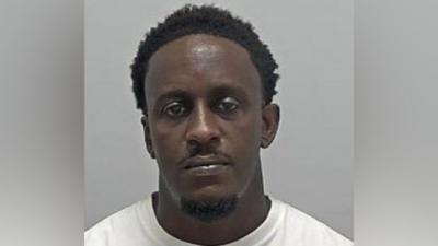 A police custody image of Joseph Gbonda's face. He has short black hair, a moustache and goatee beard, and is wearing a white T-shirt