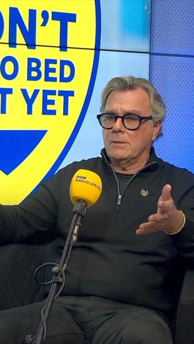 Peter Lowy sits talking into a BBC Radio Leeds branded microphone. He has grey hair, sept back from his face. He is wearing black rimmed glasses and a black zip up top.