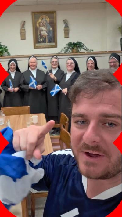 A man with brown hair and blue eyes with a short beard and moustache points at the camera. He is holding a Scotland flag and wearing a blue and white top. Behind him are nuns dressed in black and white and holding Scotland flags. They are standing in front of a wall with a religious painting hanging up beside plants.