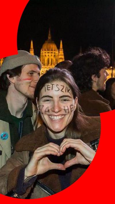 Woman with writing on her face smiling, making a heart symbol with her hands. 