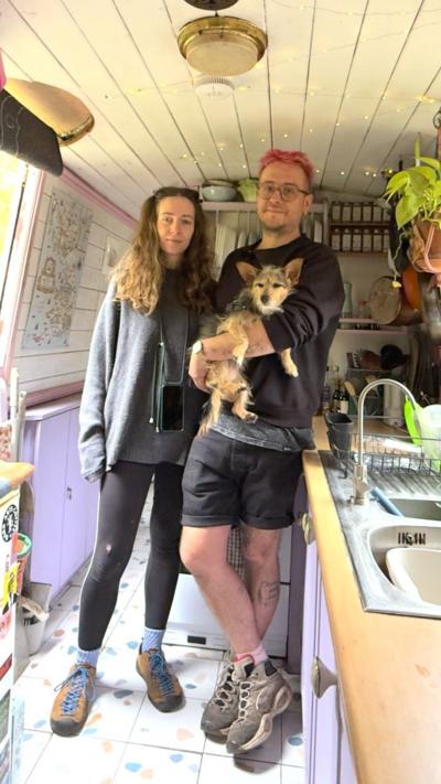A couple stand in the middle of a thin narrowboat. They are holding a small dog. 