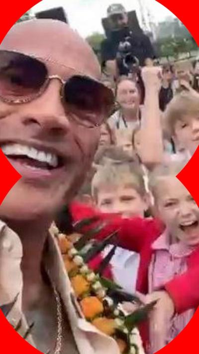 A selfie-style image taken by The Rock – a man wearing glasses and smiling. Next to and behind him is a group of schoolchildren. They are cheering, smiling and shouting. They are wearing red and white school uniforms.