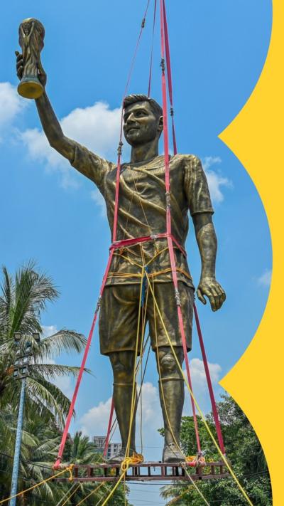 A 70 fit statue of Lionel Messi stands on a pallet in midair, secured with straps and lifting equipment