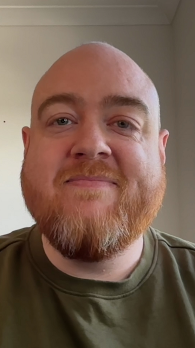 A man with no hair and a beard wears a dark top and looks at the camera