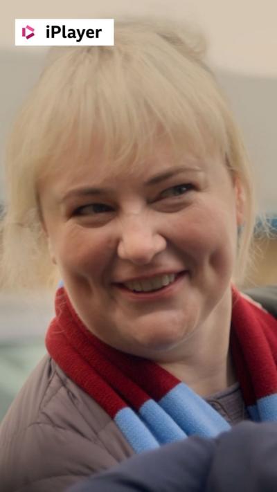 A woman with blonde hair, wearing a football scarf, looks cheekily to someone off camera.