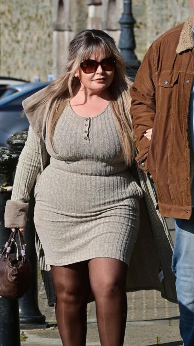 A woman wearing sunglasses and a grey dress walks with her arm linked to a man in a tan jacket