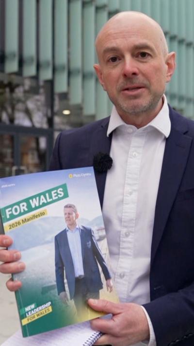 Gareth Lewis, who has a bald head, in a jacket and white shirt holding up a manifesto, with a pic of Rhun ap iorwerth and a title saying "For Wales"