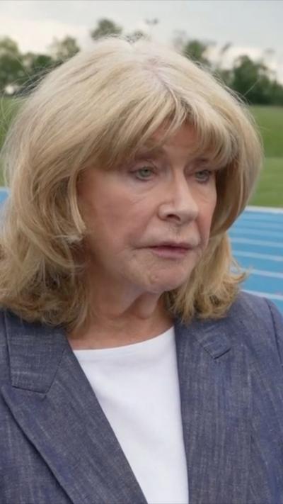 A woman with blonde hair wearing a white t-shirt and grey blazer stands in front of a blue running track