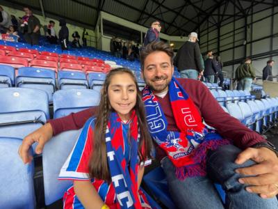 A girl and a man wear Crystal Palace football scarfs and sit in the stands. They are smiling at the camera