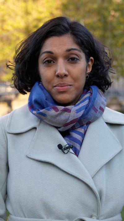 Woman wearing blue and purple scarf and beige coat. She has short black hair and is standing in front of a tree.