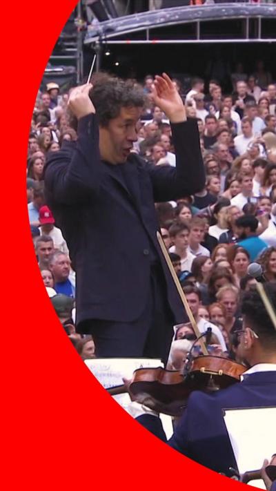 Gustavo Dudamel, in a dark blue suit, conducts an orchestra in front of a crowd