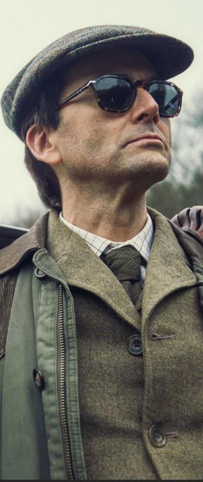 David Tennant wearing dark sunglasses and a flat cap, wearing green tweed and a green jacket in a scene from The Rivals 
