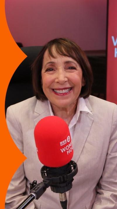 A woman with a bob haircut wearing a beige blazer sits in front of a red microphone.