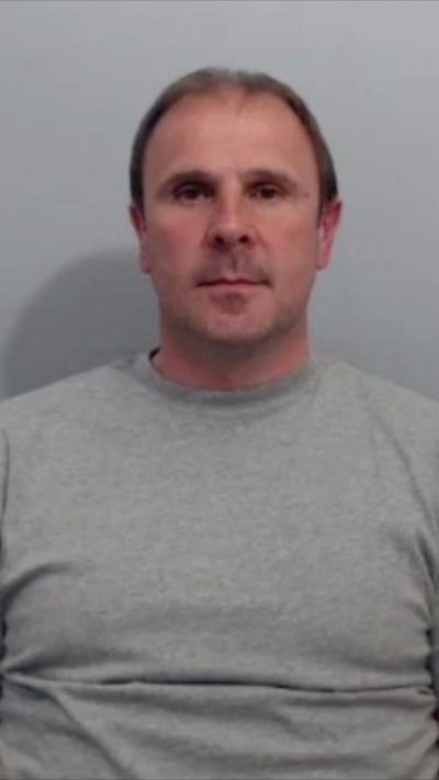 A man with short dark hair is facing the camera for a police mug shot. He is wearing a grey sweatshirt and has short, balding brown hair. He is looking directly down the lends of the camera.