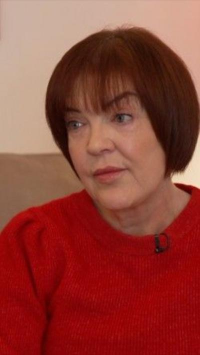 A woman with chin-length brown hair is looking off to the side of the camera. She is wearing a red jumper.