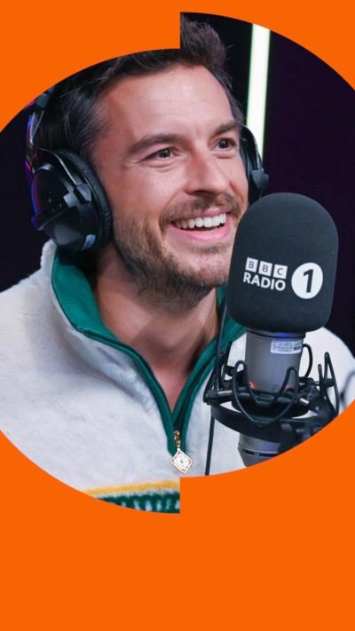 Jonathan Bailey smiles wearing headphones and behind a Radio 1 mic - he wears a white and green zip top