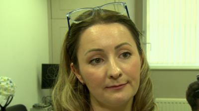 Dr Kathy Ruddy is wearing a white shirt and has long blonde hair. Her glasses are on top of her head and she has a silver pendant necklace on.