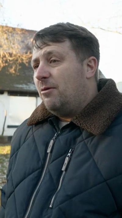 A man with brown hair wearing a blue padded coat with brown collars outside a closed pub. The building is white with The Hare & Hounds written on the wall