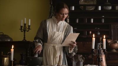 An actress playing Jane Austen. They are in a room lit by candle light and reading from a piece of paper.