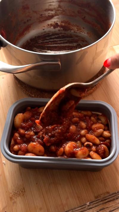 A box of the stew with a spoon ladling the beans into the box. An empty cooking pot stands behind it.