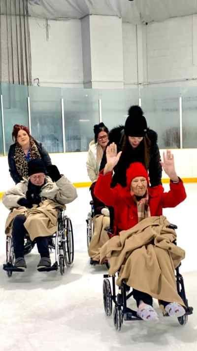 Three care home residents wearing coats and blankets sit in their wheelchairs while three people push them around an ice rink.