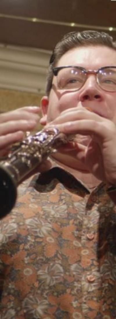 A man with dark hair and glasses in a brown, white and black flowery shirt plays the clarinet
