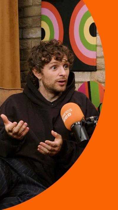 Tom Grennan speaks into an orange mic. He wears a baggy brown hoodie and has curly brown hair 