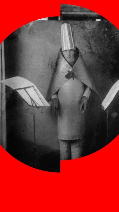 A man in a rigid cape and tall cylindrical hat stands between two music stands.