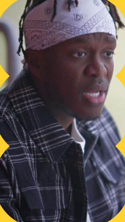 KSI wears a grey bandana and a blue checked shirt. He talks to someone off-camera.