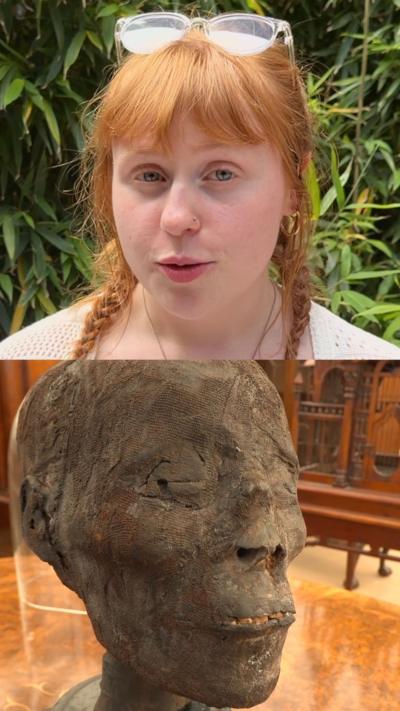 Amy Murray has glasses on top of here head, he hair is red and in pigtails. Behind here is a bush.
Below here is a mummified head.  