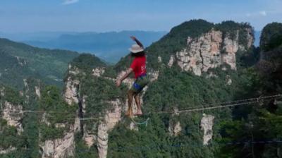 A man wearing a red t-shirt and hat balances on a slackline high above a gorge in China. In the background, tall rock faces covered in green trees reach far into the distance.
