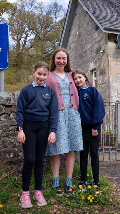Pupils Molly and Shannon stand with their teacher Holly outside Duror Primary, a small stone building.