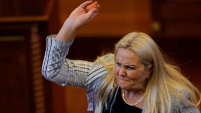 Blond woman wearing white and black blazer lifting right arm and holding egg 