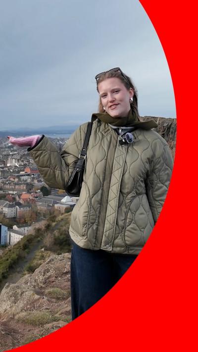 A woman in a green jacket gestures to a view of Edinburgh behind her 
