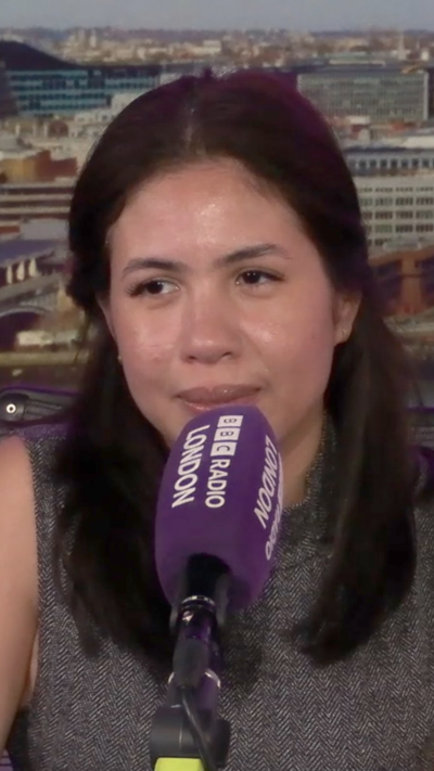 A woman speaks into a microphone against a backdrop of a city skyline and purple.
