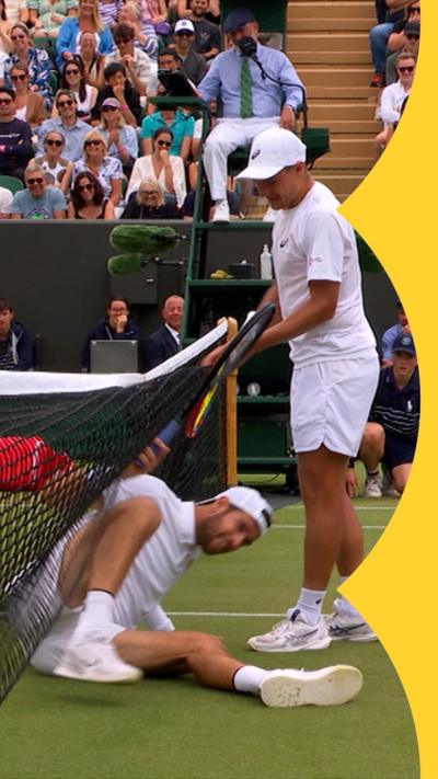 Male tennis player wearing all white laying on side underneath net while another tennis player, standing, moves the net with his raquet to help him up
