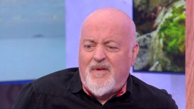 A man, Bill Bailey, looks confused; he is bald, has a grey beard and is wearing a black shirt