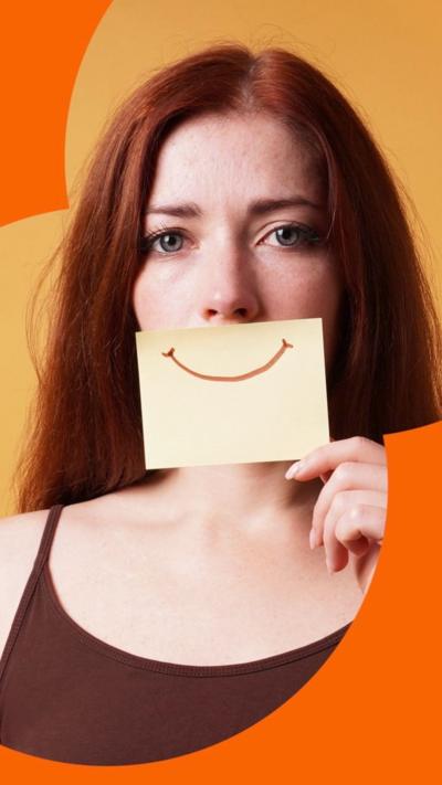 A woman with long hair wearing a strappy brown top, holding a piece of paper with a smile drawn over it, over her mouth.
