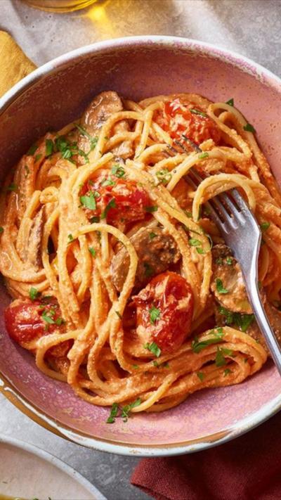 Top down view of a pink bowl containing red pepper pasta with a fork in it. A red napkin sits by the side.