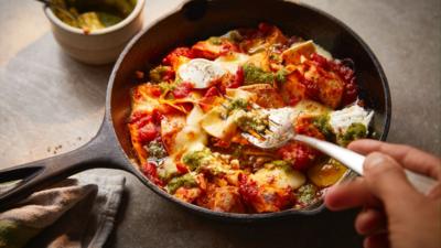 A one handled pan with chicken lasagne in. A hand holding a fork can also be seen. 