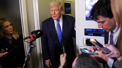 Donald Trump wearing a suit and blue tie speaks to reporters holding up microphones and phones through an open door.