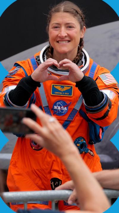 A woman in an orange spacesuit smiles as she makes a heart shape with her hands 