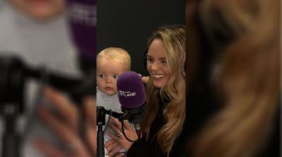 A woman smiles holding her baby. They are in a radio studio with a purple mic in front of them.